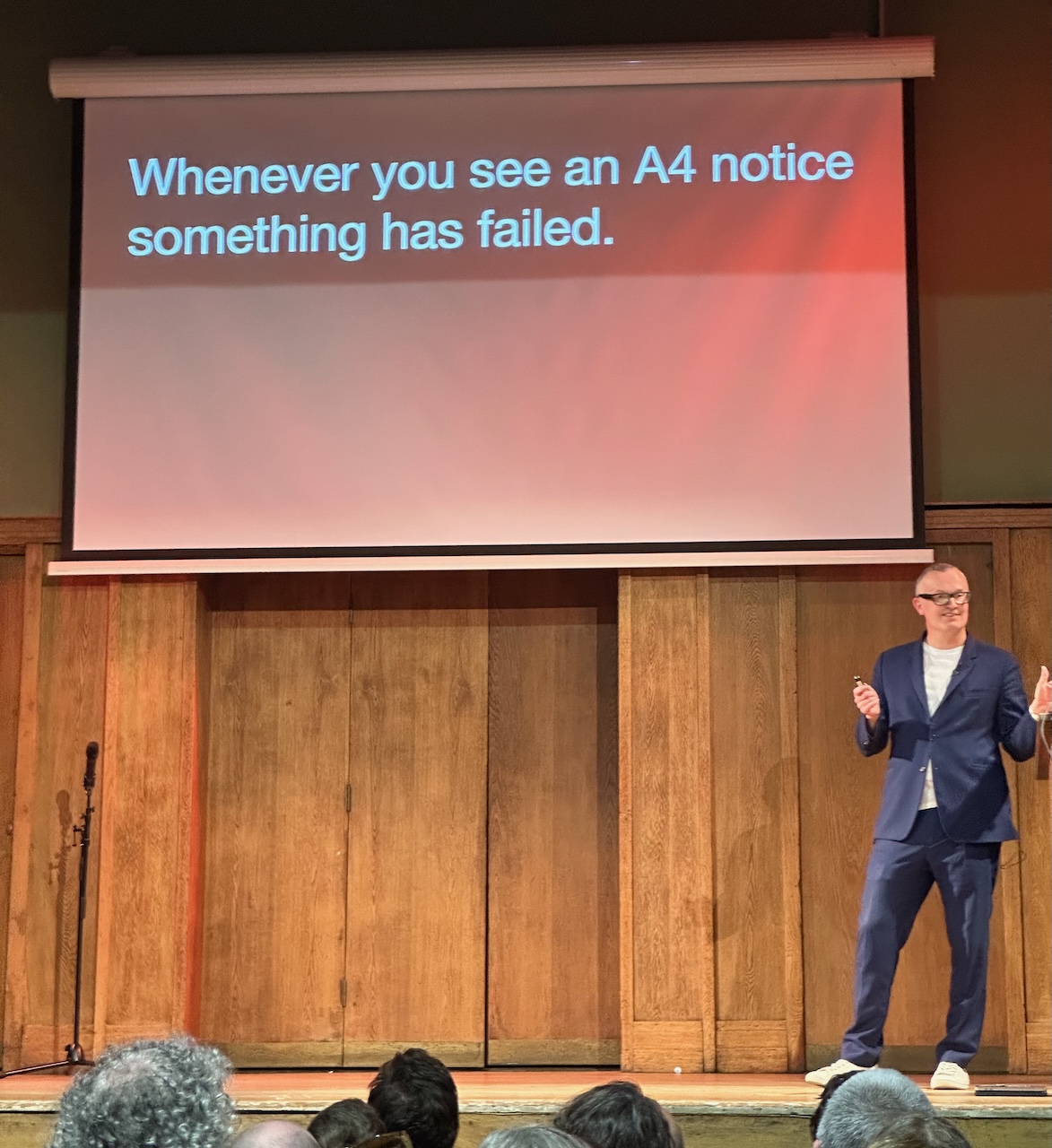 A man in a blue suit standing on a wooden stage in front of a projected slide which just has the test “whenever you see an A4 notice, something has failed’