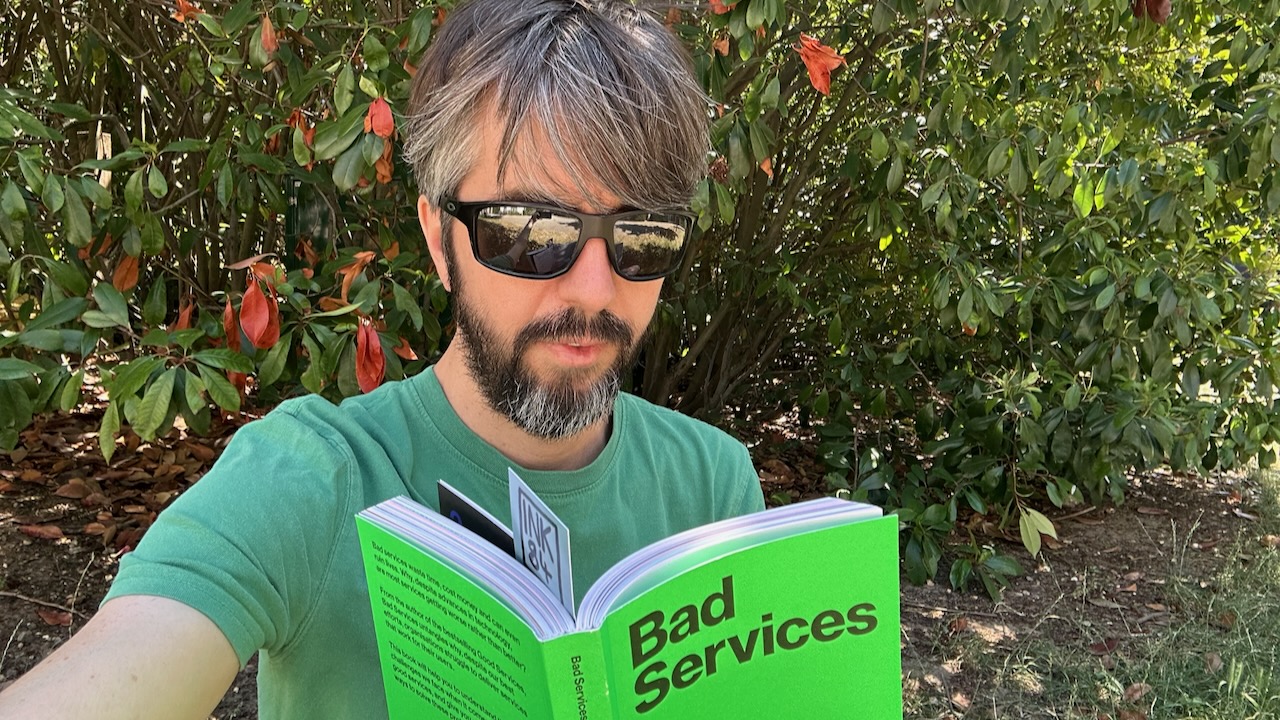 Photo of me sat outside reading a bright green book with the title 'Bad Services'