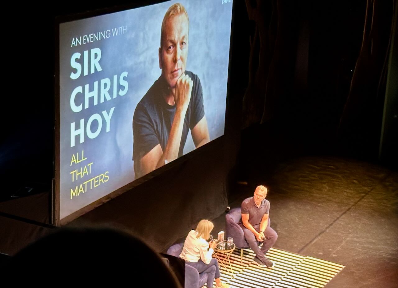 Photo of a stage lit by strong spotlights, on which sit a man and a woman, and behind them a large screen saying ‘An evening with Sir Chris Hoy’ – All that matters