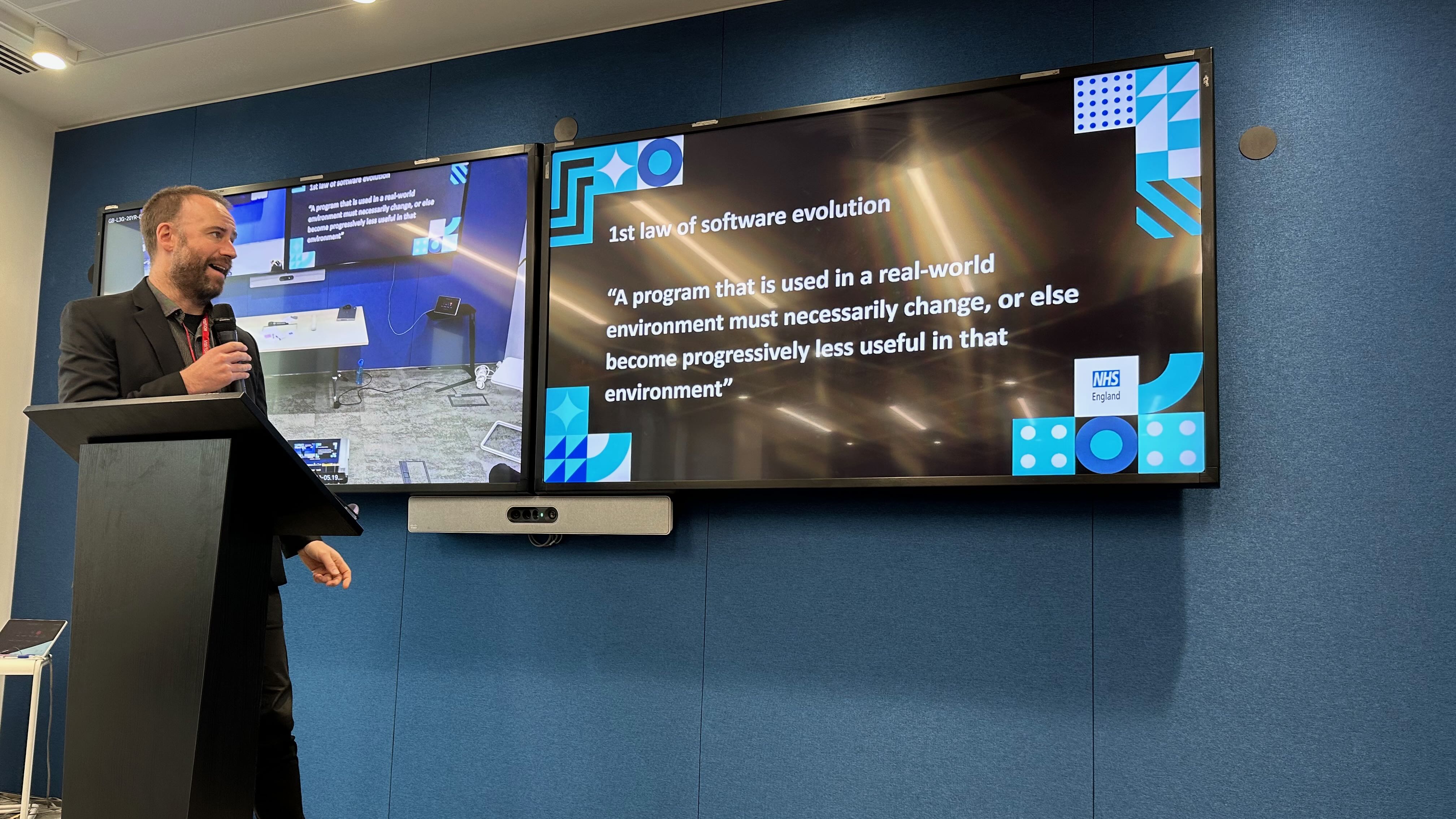 Photo of a man presenting in front of a screen which has the the text: 1st law of software evolution: “A program that is used in a real-world environment must necessarily change, or else become progressively less useful in that environment.”