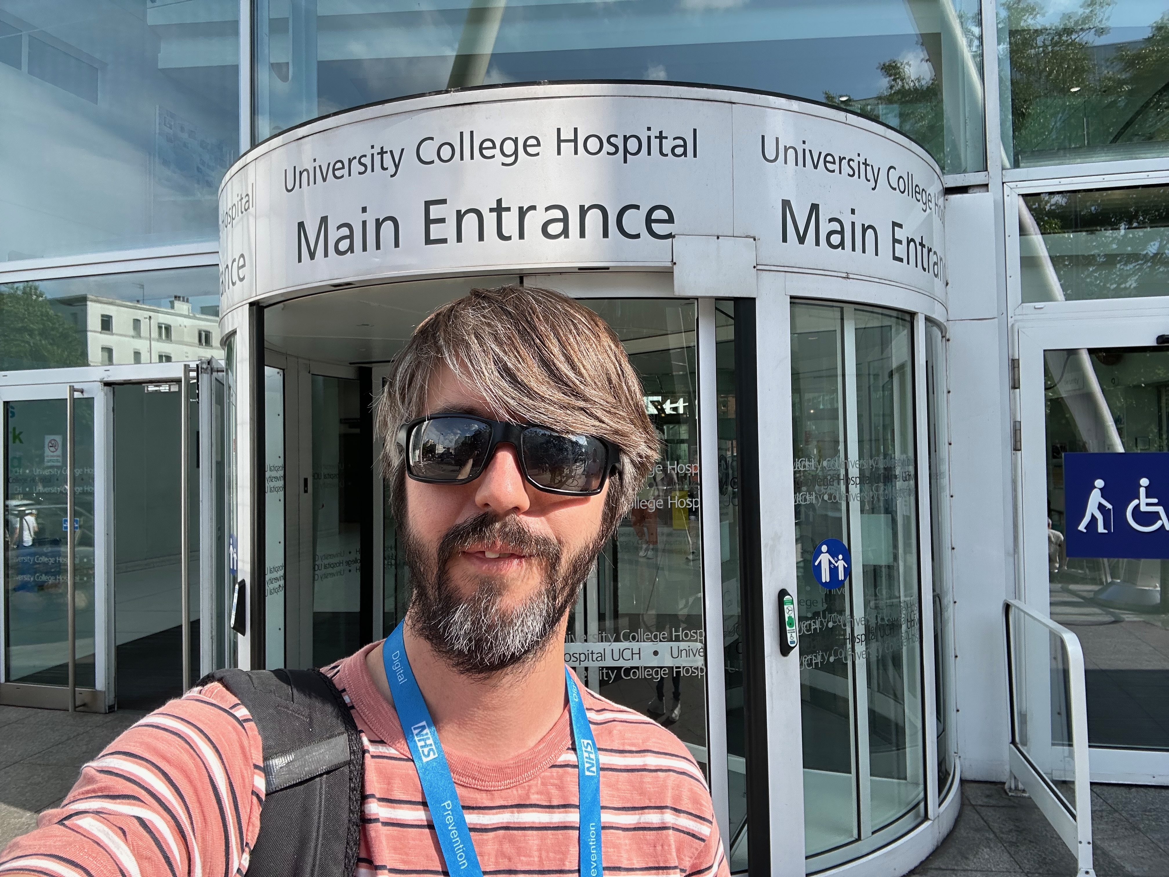 Photo of me with a blue NHS lanyard on, standing outside a revolving door with the sign above it reading ‘University College Hospital main entrance’