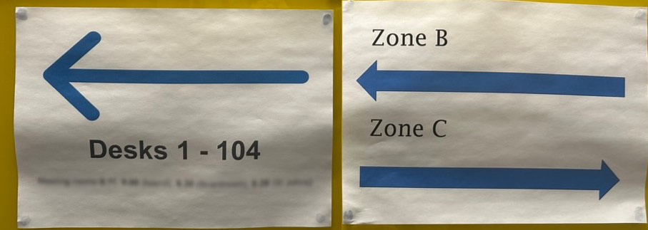 Photo showing two signs side-by-side, one with pointing at desks 1 to 104 and the other to zones B and C. Each one uses completely different blue arrows.