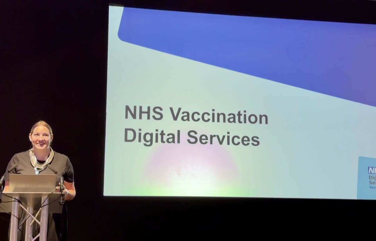 Photo of a Helena in front of a lectern and screen with the words NHS Vaccination Digital Services on it