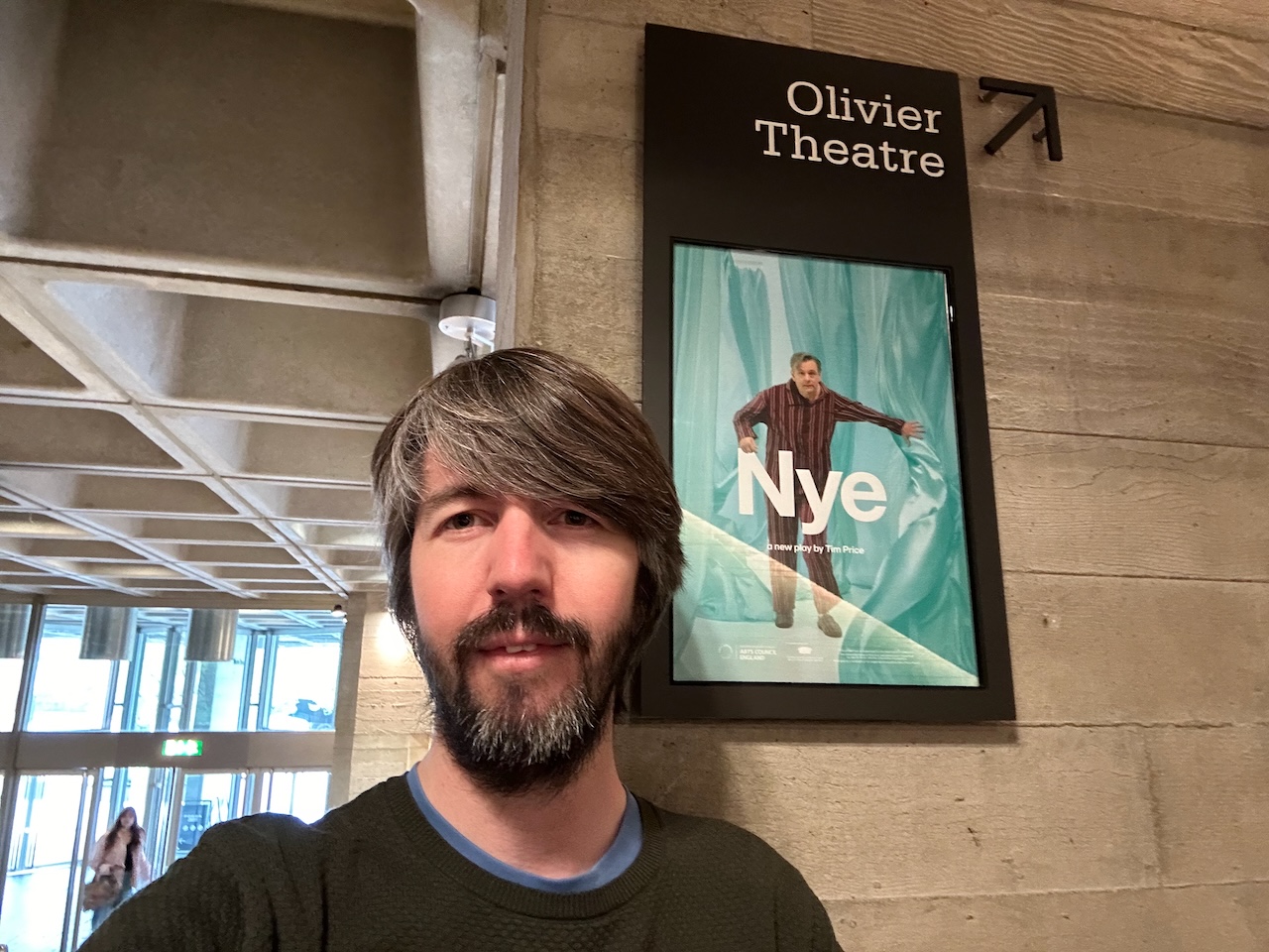 Photo of me standing against a brutalist concrete wall next to a post for the play Nye