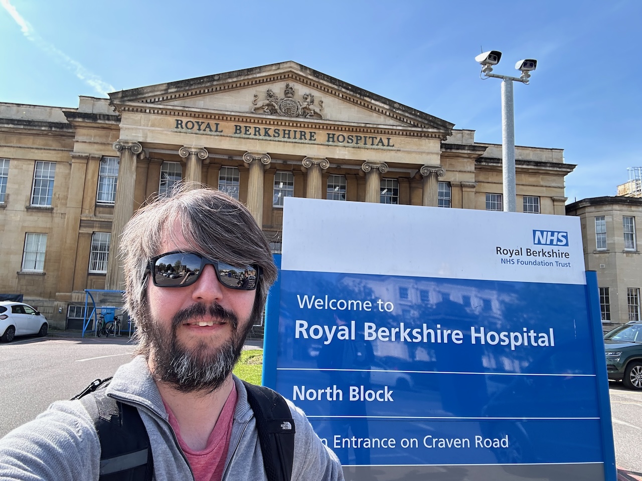 Photo of me standing in front of an NHS sign saying Welcome to the Royal Berkshire Hospital, behind which is an old stone portico with the same name above the columns.