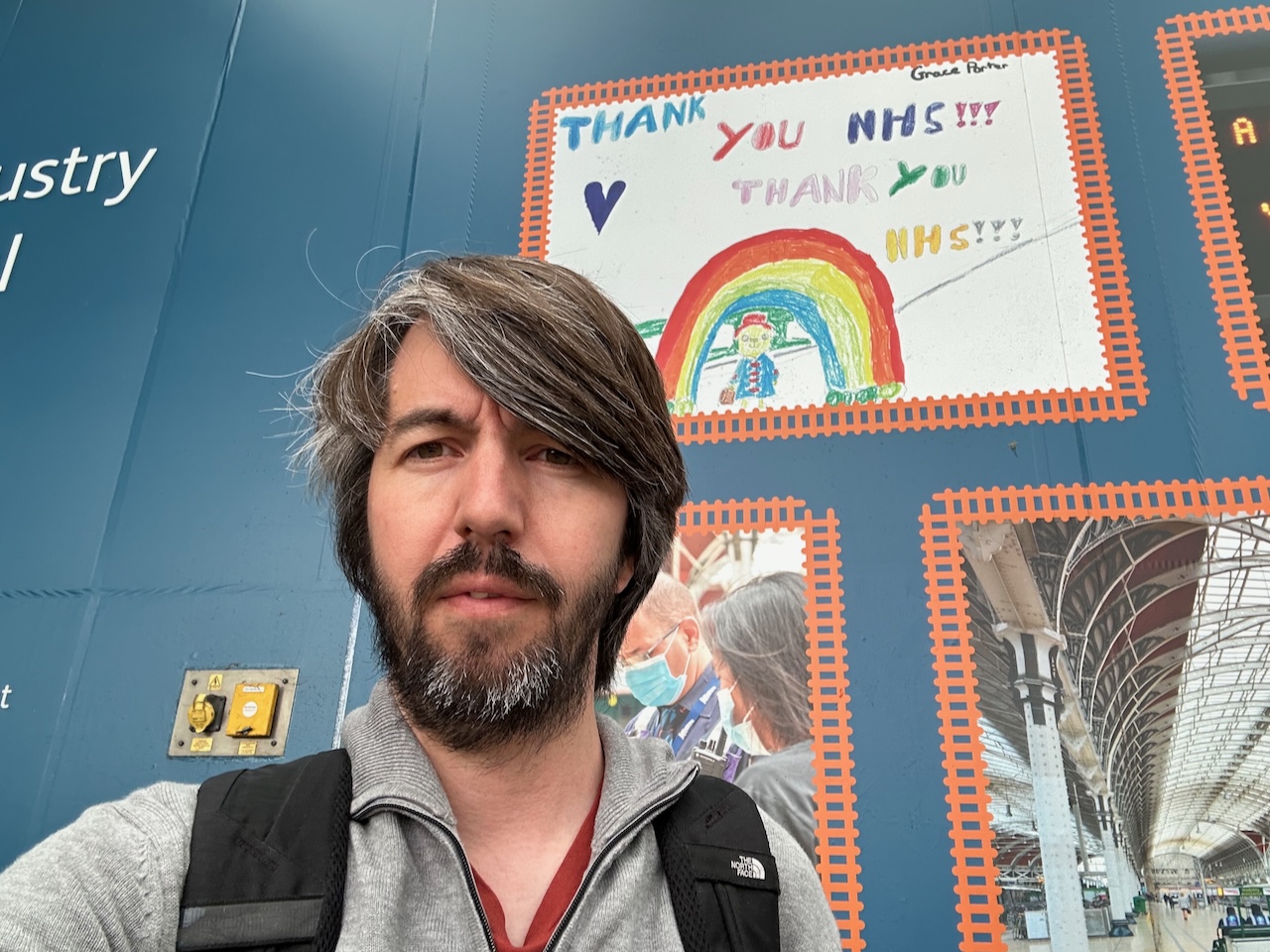 Photo of me standing in front of a colourful child’s drawing of rainbow with the word ‘Thank you NHS!!’ printed on some hoarding at a train station