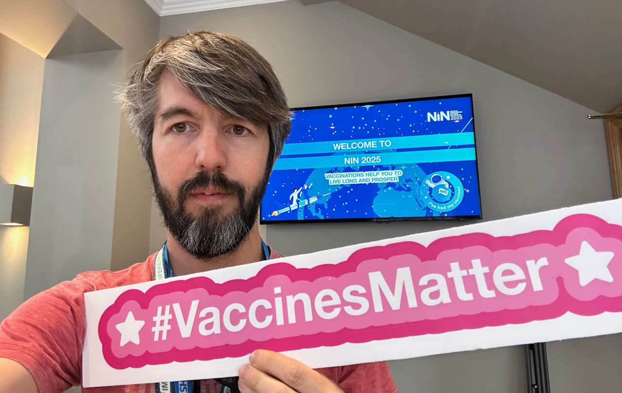 Photo of me in front of a TV screen saying Welcome to NIN 2025 and holding a sign up saying #VaccinesMatter