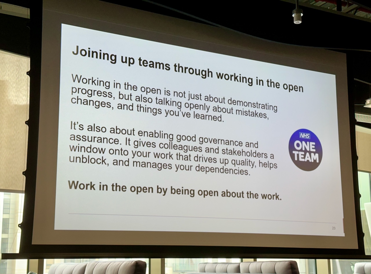 Photo of a projector screen showing a slide with the text “Joining up teams through working in the open. Working in the open is not just about demonstrating progress, but also talking openly about mistakes, changes, and things you’ve learnt. It’s also about enabling good governance and assurance. It gives colleagues and stakesholders a window onto your work that drives up quality, helps unblock, and manages your dependenceis. Work in the open by being open about the work.”